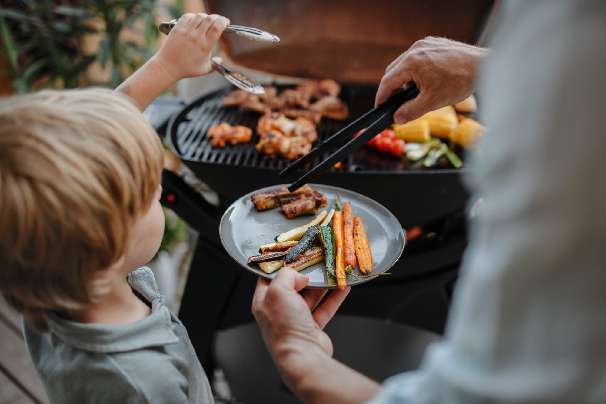unrecognizable father with little son grilling ribs and vegetable on grill during family summer garden party close-up